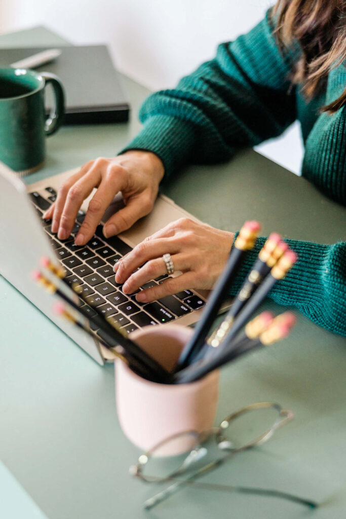 Woman at laptop typing with cup and pencils on side